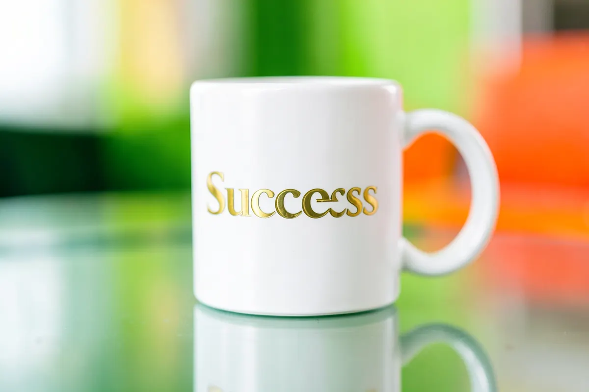 Close-up de uma caneca branca com a palavra "Sucesso" escrita em letras douradas elegantes, sobre uma superfície espelhada.