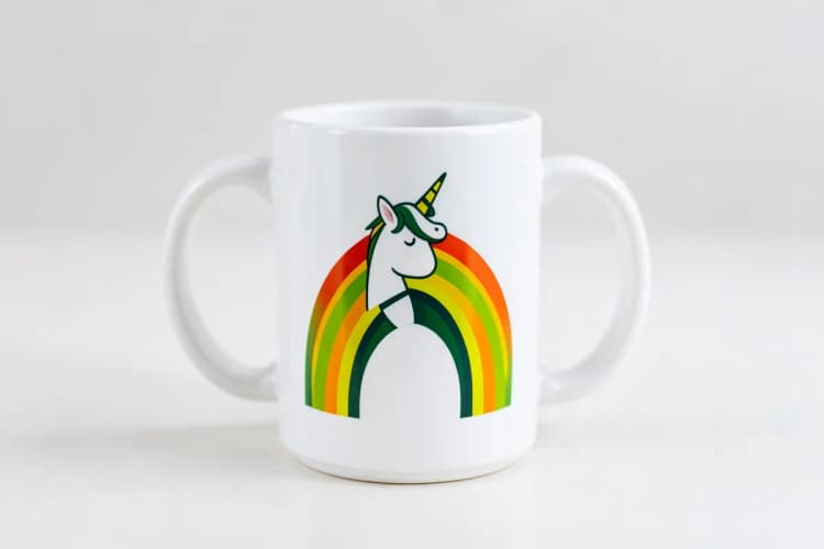 Caneca branca com design de um unicórnio sorridente e arco-íris, em plano detalhe, sobre superfície clara.