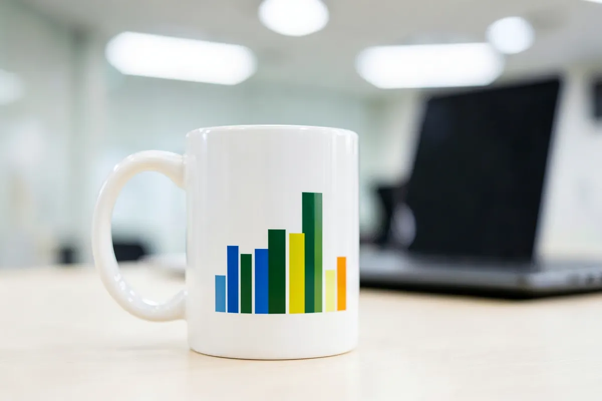 Caneca de cerâmica branca com estampa de um gráfico de barras ascendente em tons de azul, em plano médio, sobre uma mesa de escritório com um notebook ao fundo.