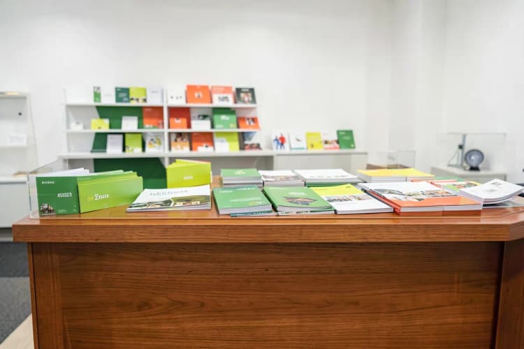 Balcão de atendimento de uma gráfica com um atendente sorridente e materiais impressos organizados ao fundo.