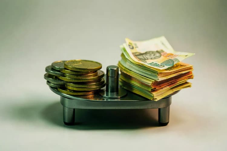 Uma pequena balança de precisão com moedas de diferentes valores em um prato e notas de dinheiro dobradas no outro, simbolizando custo.