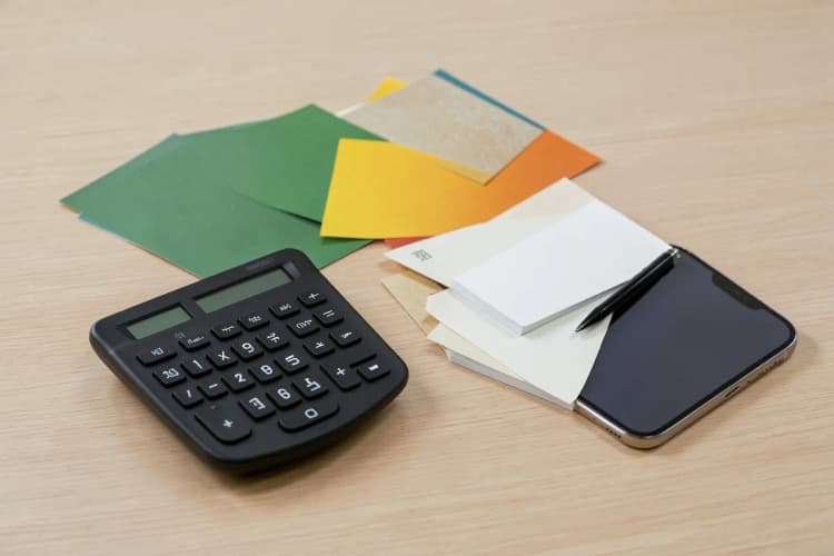 Calculadora e bloco de notas com caneta sobre uma mesa, ao lado de amostras de impressos e um smartphone.